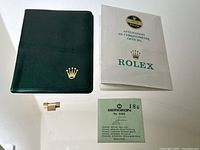 All lot items on white surface: gold link, green vinyl holder, folded Rolex booklet, Bergeon packet
