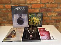 Five hardcover coffee table books arranged on a white table against a brick wall: Lalique, Tiffany, Carl Fabergé, Famous Diamonds, and Baccarat.