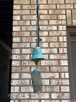 Full view of the bronze bell wind chime hanging from a hook on a brick wall, showing the blue-green patina on the bell and the metal hanger.