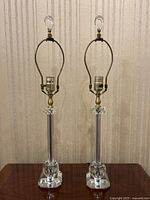 Front view of two clear lucite lamps showing octagonal bases, cylindrical lucite columns, brass finishing near the sockets, and lucite finials.