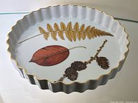 Top view of the white scalloped ceramic pie plate showing detailed botanical motif of brown pine cones, red leaf and golden fern leaf with a gold trimmed rim.