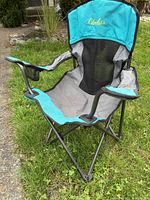 Front angled view of teal Cabela's folding kids chair on grass, showing overall seat, armrests, and mesh back.