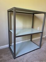 Full front angle view of the glass and metal shelving unit showing three shelves and metal frame against a wall.