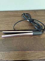 Electric hair straightener iron with rose gold and black color and cord coiled beside it, shown from the side revealing the plates and hinge.