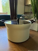 Ivory-colored cast iron stockpot with glass lid on wooden table. Pot has sturdy handles and black handle on lid.