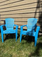 Two blue plastic kids outdoor chairs shown side by side on grass in front of siding wall.
