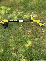 Full side view of Ryobi battery-powered lawn edger and trimmer laying on grass showing the full length and details of the shaft, cutting guard, and rear battery housing
