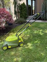 Electric green lawn mower shown from side in grass with garden background.
