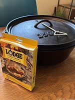Photo showing the Lodge box and the cast iron Dutch oven with the lid on and the metal lid lifter hook resting on top.