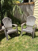 Two brown plastic Adirondack-style outdoor chairs sitting on grass near a wooden fence and siding, shown together.