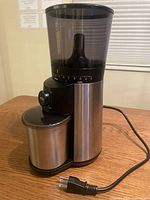Front and top view showing the black tinted bean hopper, grind size selector dial, and stainless steel ground coffee container.