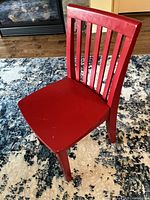 Red solid wood child's chair from Pottery Barn Kids shown on rug with fireplace background; front angle showing scratches on seat surface.