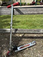 Photo of the Razor Classic scooter standing upright on the ground outdoors, showing the folding mechanism, red handle grips, and one red front wheel.