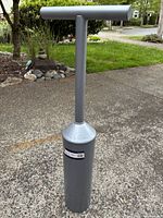Photo of the stainless steel clam digging tool standing upright on a concrete surface outdoors with greenery in the background. Shows full tool with T-handle and cylindrical digging end.