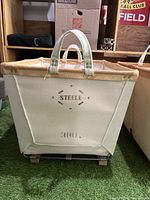 Full view of the canvas basket showing natural colored canvas body, leather rim, canvas handles, and 'STEELE 380' markings.