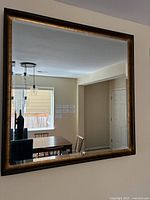 Front view of the square wall mirror hanging on a beige wall, showing the black and rustic gold frame with minor wear and the reflected interior room details.