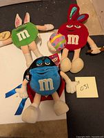 Three M&M character stuffed toys including red and green bunny versions and a blue M&M with cape