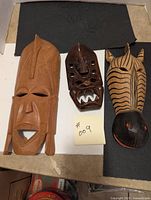 Front view of three wooden masks including a large brown mask, a smaller brown mask with white teeth, and a zebra-striped mask with red eyes