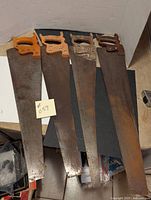 Four hand saws with wooden handles placed side by side, showing varying degrees of rust and different handle colors and styles.