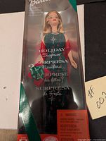 Front view of Holiday Surprise Barbie Doll in original sealed retail box, showing doll in festive green dress with red wrap, clear window packaging with green border and orange warning label.