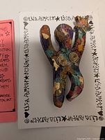 Close-up photo of Lisa Parker branded card with colorful, stylized female figure brooch that has arms raised, decorated with gold leaf and spiral details.