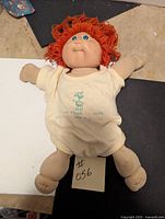 Full view of red-haired Cabbage Patch doll lying down, in cream onesie with text and graphic.