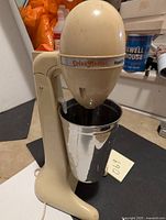 Front view photo of the Drink Master mixer, showing beige housing, metal mixing cup, and branding.