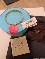 Kate Spade gold-tone bangle bracelet displayed on top of its turquoise round box with original tag and a brown fabric pouch beside it.