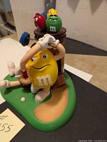 Side view showing yellow M&M character mid-golf swing with red and green M&Ms and golf bag on green base.
