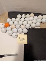 36 assorted golf balls mostly white with one orange, laid out on a black and white surface
