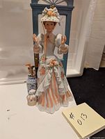 Front view of Mrs. Albee porcelain doll in white and peach dress with floral applique, holding the hat strings and standing upright next to golf club accessory.