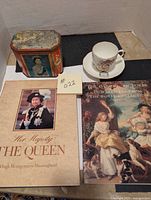 Top view of the lot items including a teacup and saucer set, vintage tin, and two hardcover books about Queen Elizabeth II and the Royal Collection.