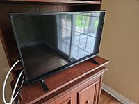 Black framed 32 inch RCA LED television placed on a wooden cabinet. TV screen is off showing reflections of window and room.