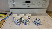 Five blue flower patterned mugs and one miniature blue floral teapot placed on a stovetop.