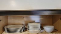 Image showing a cupboard shelf with 7 white ceramic dinner plates stacked on the left and 6 white ceramic bowls stacked on the right.