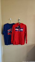 Photo showing a red Buffalo Bills XL hooded sweatshirt and a blue Buffalo Bills XL short sleeve t-shirt hung on hangers against a beige wall.