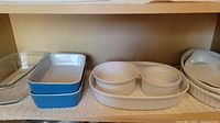 Photo shows a variety of casserole dishes and ramekins arranged on a shelf. Includes clear glass rectangular dish, blue and white ceramic rectangular dishes, two white round casserole dishes stacked, two white ribbed ceramic ramekins on a white oval dish, and a white oval ceramic dish.