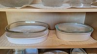 Two large white oval Pyrex casserole dishes with clear glass lids, placed on a shelf.
