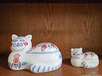 Two white ceramic cats with decorative hand-painted floral patterns, one larger with a removable lid, one smaller open container.