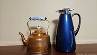 Photo of hammered copper kettle and blue thermal coffee carafe side by side on wooden surface against plain wall.