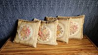 Front view of four square floral tapestry cushions arranged on a wooden surface against a dark patterned wall.