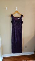 Front view of purple sleeveless sequin evening dress on a wooden hanger against beige wall.