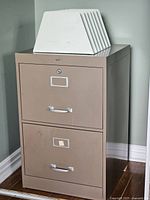 Photo of a beige two-drawer metal file cabinet with lock and handles. A white metal file holder sorter with multiple slots is placed on top.