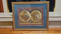 Framed antique style world map print measuring approximately 20x24 inches. Gold color ornate carved frame surrounds two hemisphere map print with vintage decorative imagery. Blue mats inside the frame.