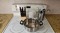 Image showing a white stove top with a large stainless steel pot, a wooden knife block with knives, a transparent plastic pitcher, and a stainless steel utensil holder with several kitchen utensils.