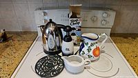 Photo of kitchen items on stovetop including metal electric kettle, Slap Chop chopper in box, ceramic pitcher, gravy bowl, and black metal trivet.
