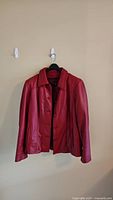 Photo of red leather jacket by Danier sized large, hanging on a wall with front buttons visible.