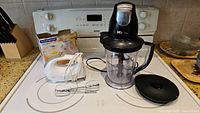 Shows the Ninja food chopper blender with lid, and white electric hand mixer with beaters on a kitchen stove.