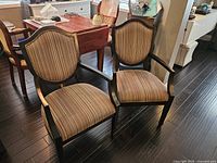 Two dark wood framed occasional chairs with padded striped upholstery on seats and backs, placed on dark wood floor near wooden table.