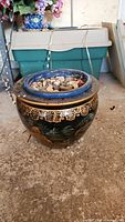 Single photo showing a round ceramic planter with a blue rim and black body with gold detailing filled with small natural pebbles.